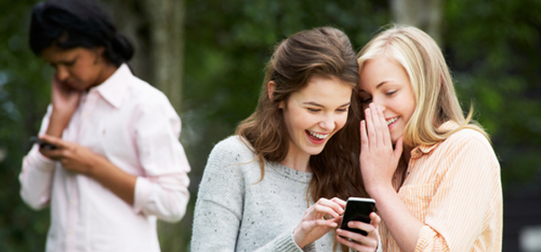 An upset teen girl looks at her phone. A group of girls are standing behind her, looking at her and laughing.