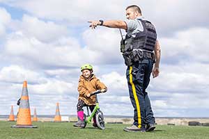 A young child wearing pink rainboots and a green helmet sits on a green bicycle looking toward the left. A male RCMP officer points in the direction she is looking. Orange cones are on the grass beside them.