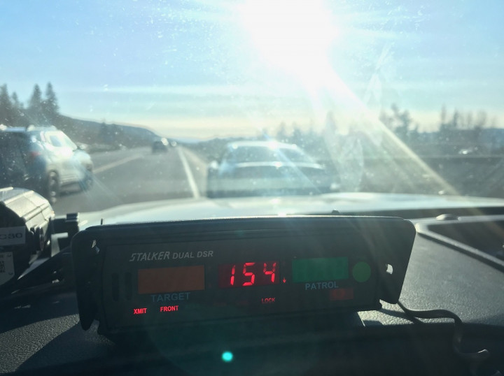 The inside of a police vehicle shows a radar unit mounted on the dash with a speed displayed as 154 km/hr. A car is pulled over on the shoulder of the highway during a sunny day in front of the police vehicle.