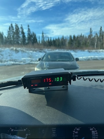 A picture is taken from the inside of a police vehicle. A radar unit on the dash displays a speed of 175 km/h. A parked car faces the police vehicle head on.