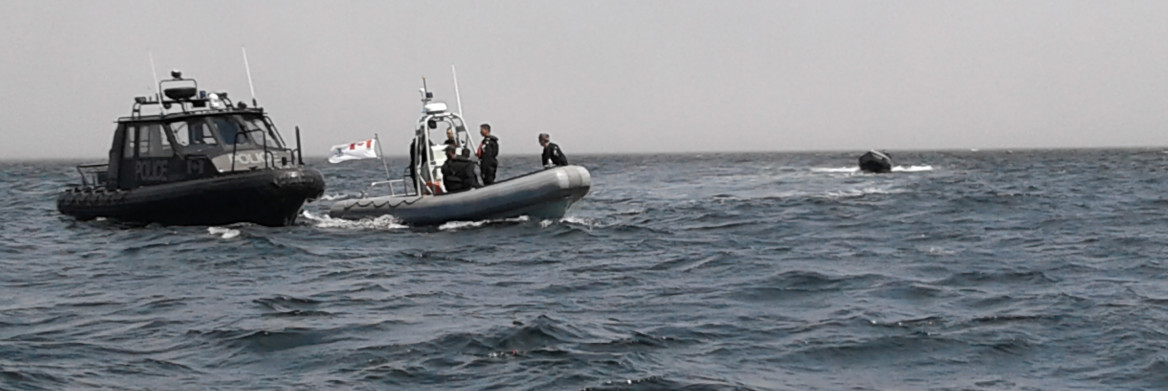 Trois policiers dans une embarcation abordée par une autre embarcation en mer. 