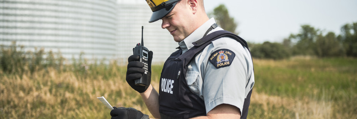 Male police officer stands in a field while holding mobile radio and looking at notes, with silos in background.