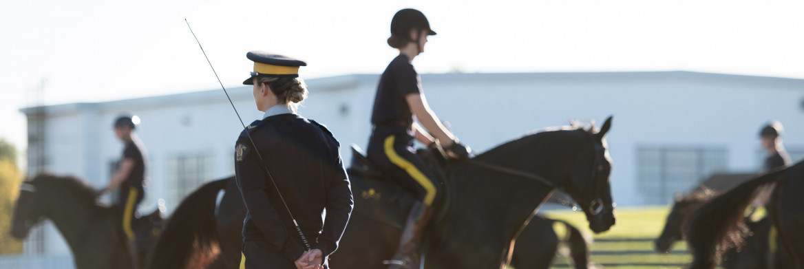 A female RCMP officer stands in a riding ring, holding a crop. She watches other officers ride their horses.