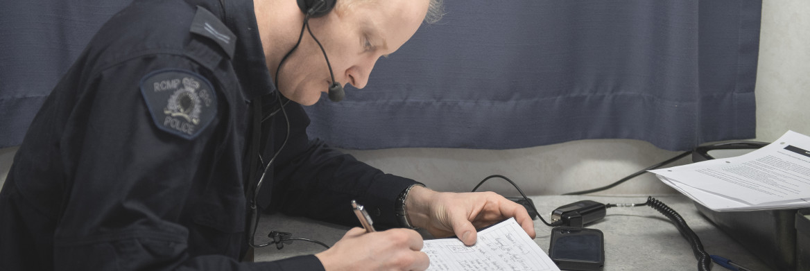 A male RCMP officer wearing headphones sits at a desk writing notes. 