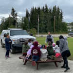 A group of small school children sitting at a picnic table learning from teachers and police officers.