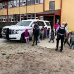 A group of school children drawing on a police vehicle with the assistance of police officers using dry erase markers as part of the Colouring a Police Vehicle event.
