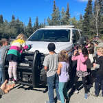 A group of school children colourfully drawing on a police vehicle using dry erase markers as part of the Colouring a Police Vehicle event as community RCMP officers and teachers supervise them.