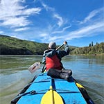 The back of a youth at the front of a two-person canoe paddling along the river under beautiful blue skies