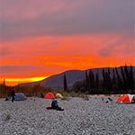 Red sunset skies and multiple tents set up along the gravel shores of the river