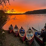 Empty canoes tied along the shore of a river under a red sunset