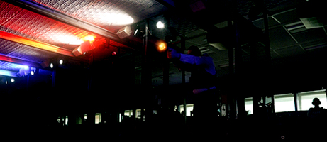 A cadet shoots their pistol from a stall in a dark range while red and blue lights flash above them.