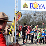 A large group of children line up at a race start line. A Mountie cheers from the side!
