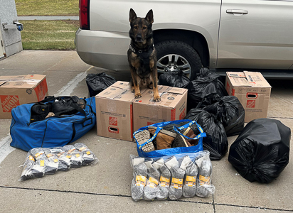 A German shepherd police dog is sitting atop two boxes; the dog is surrounded by bags of boots and packages of new socks.