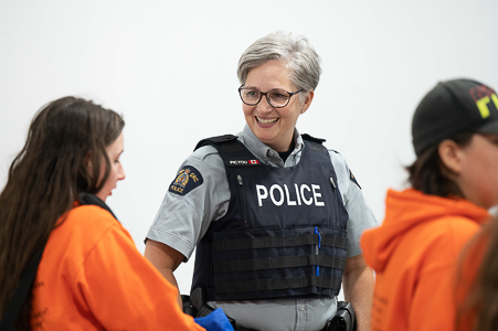 A female police officer, who’s wearing glasses, is smiling at a young girl; the girl, who’s wearing an orange top, has long dark hair.