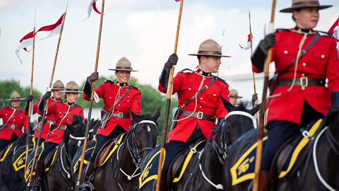 Several RCMP officers, carrying red and white flags, are riding black horses while wearing red serge.