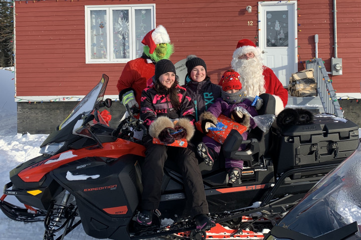 From Makkovik, Newfoundland - Three children sitting on a snowmobile with wrapped toys in their hands. Santa and the Grinch standing behind them.