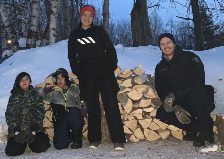 Un policier de la GRC, une enseignante et 2 garçons devant une pile de bois de chauffage coupé 