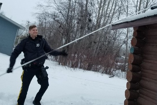 An RCMP officer (male) clears snow off a roof with a long handled snow rake