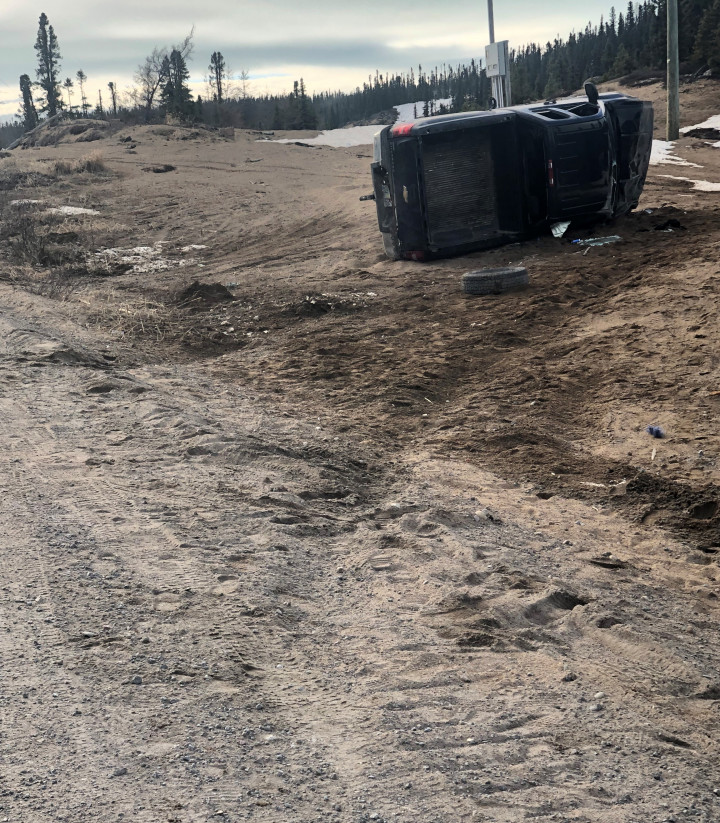 Tire tracks in the dirt lead to a pickup truck on its side. One tire is in front of the vehicle, with trees in and a dark sky the background. 