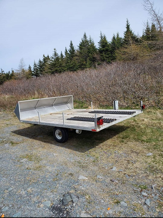 A small grey utility trailer is parked on the gravel beside a wooded area.