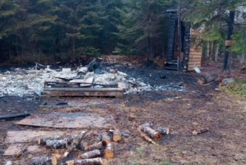 The burnt remains of a warm-up shelter after being destroyed by fire. The ground is black with burnt debris. An outhouse which was located behind the warm-up shelter is partially burnt. 