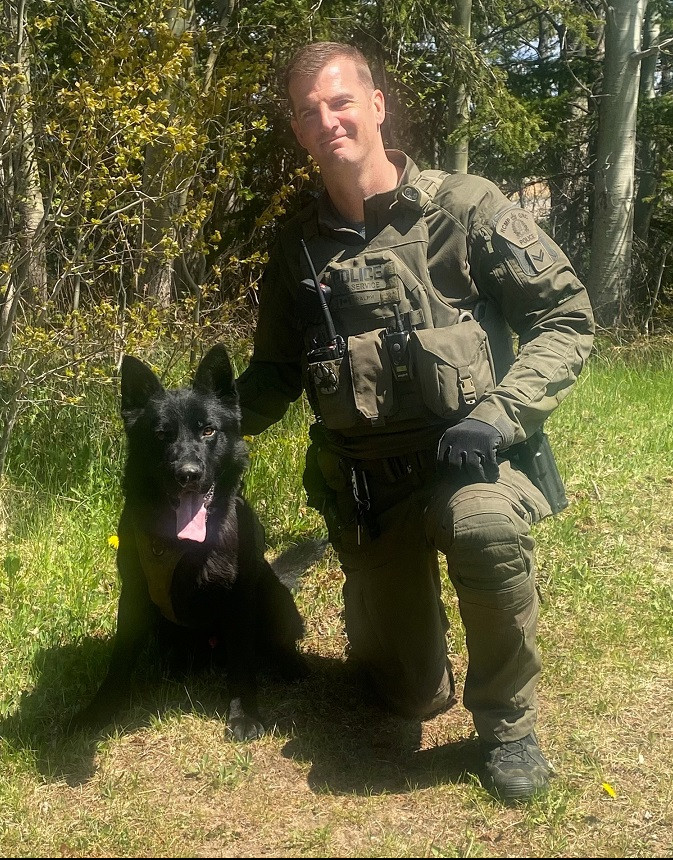 A RCMP Police Dog Services Handler is kneeling on the grass beside a black German Shepherd Police Service Dog in front of a wooded area.