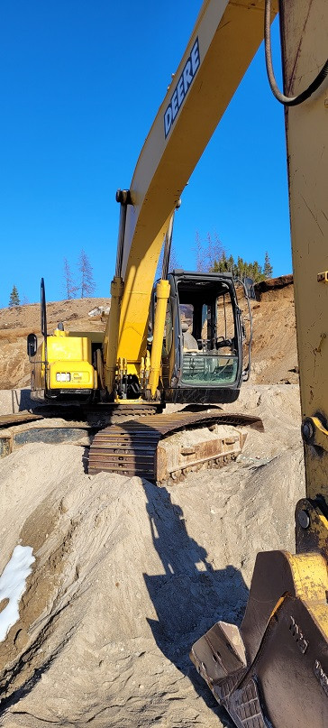A yellow John Deere excavator is parked in a sand pit with the bucket down on the ground. A blue sky can be seen in the background.