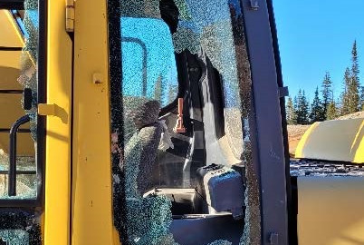 The cab of a yellow John Deere excavator is pictured with multiple windows smashed. A blue sky can be seen in the background.