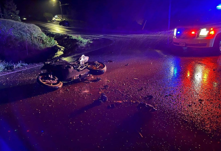 A motorcycle lies on its side in the roadway at night with debris scattered across the road. A marked police vehicle with its lights activated is pictured in the top left corner.