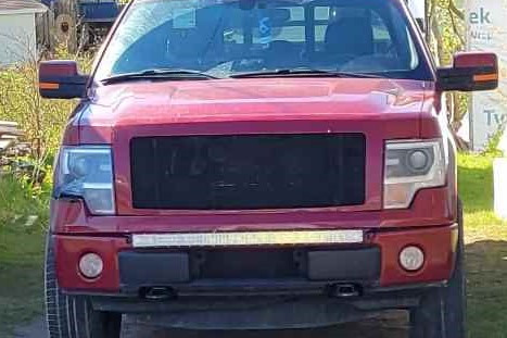 A red pickup truck is shown front on, with the front grill blacked out. A trailer is attached on the back of the truck and sheds and a house are seen in the background.