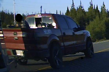 A red pickup truck with a trailer hitch on a road, driving on the yellow line, with a man in the back pan of the truck. 