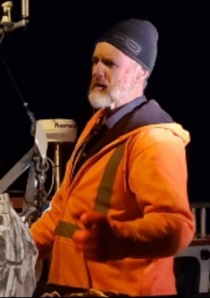 Un homme dans la soixantaine est debout, avec le bras gauche levé. Il a une barbe grise, et il porte un manteau et des gants de travail orange, et un chapeau foncé.