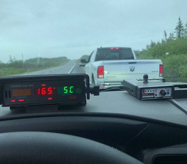Police radar on the inside dash of a police vehicle displays a speed of 165 km/h. A white Dodge pick-up truck is stopped on the side of the highway