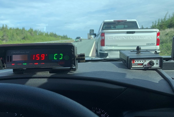 Police radar on the inside dash of a police vehicle displays a speed of 159 km/h. A white Chevrolet pick-up truck is stopped on the side of the highway.