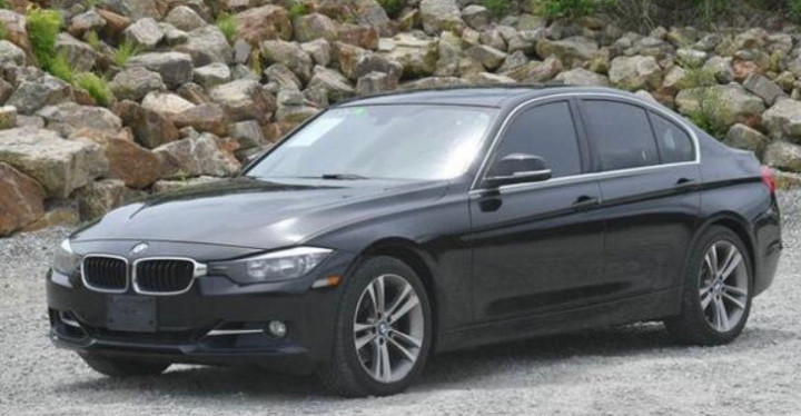 A four-door black BMW sedan is parked on gravel in front of a rocky background.