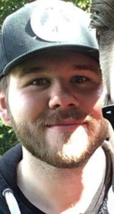 A man is wearing a dark colored ball cap and has brown facial hair.
