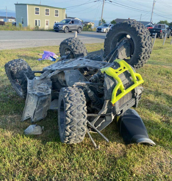 A grey and fluorescent green ATV is seen upside down with the seat detached laying on a grassy field near a roadside.
