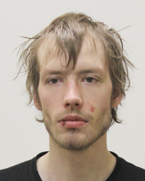 A passport-style picture of a man with short light brown hair, green eyes and facial hair. He is wearing a black T-shirt. 