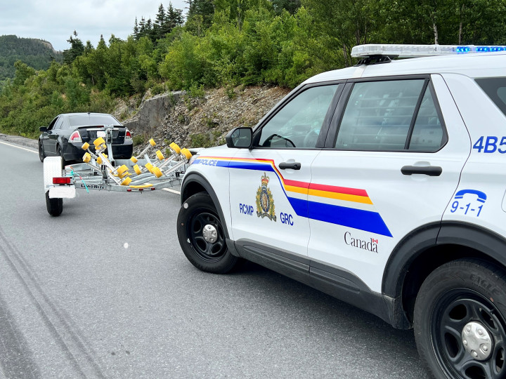 A black four-door car with a boat trailer attached is pulled to the side of the road, in front of a marked RCMP vehicle. 