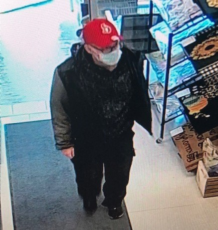 An individual wearing dark-rimmed glasses, a red ball cap and a mask enters a store.