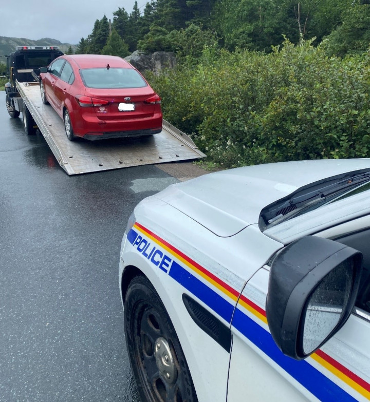 A red four-door vehicle sits on the back of a tow truck in front of a marked RCMP police vehicle on the side of a road. 