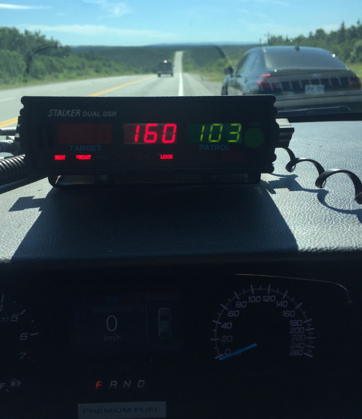 Le radar d'une voiture de patrouille indique une vitesse de 160 km/h, en rouge, et de 103 km/h, en vert. Par le pare brise, on voit la route et une berline grise rangée sur le côté de la route lors d'une journée ensoleillée.