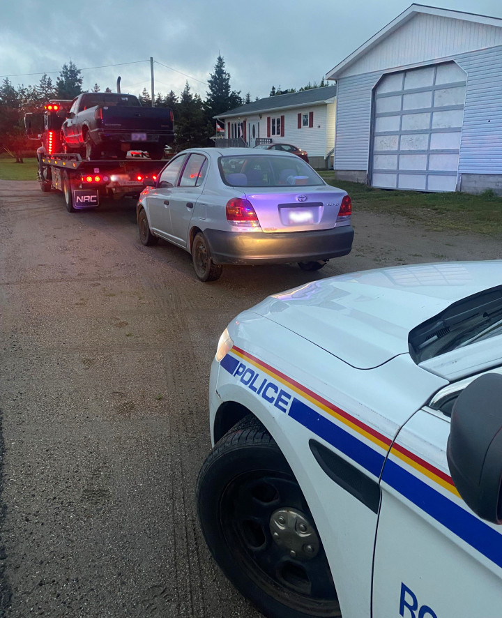 A dark-coloured pickup truck sits on the back of a tow truck with red and orange lights activated. Behind the tow truck is a grey four-door Echo car and a marked RCMP police vehicle with its headlights on. 