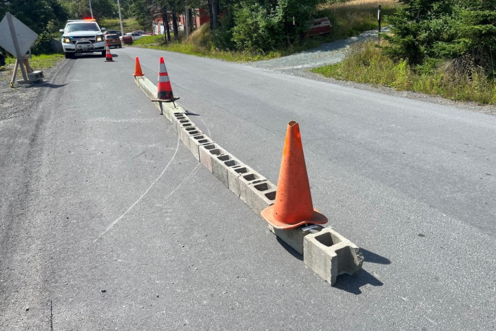 De nombreux blocs de ciment forment une barrière sur une rue résidentielle à Spaniard's Bay, en plein jour. Des cônes ont été déposés sur la barrière et un véhicule de police de la GRC stationné est visible en arrière plan.
