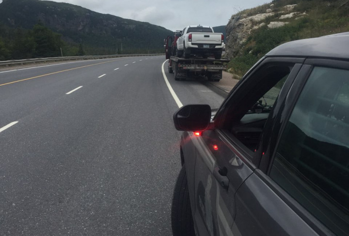 La photo montre le côté conducteur d'une voiture de police non identifiée et, un peu plus loin devant, sur la route, une camionnette blanche sur la plateforme d'une dépanneuse, sous une journée à ciel couvert.