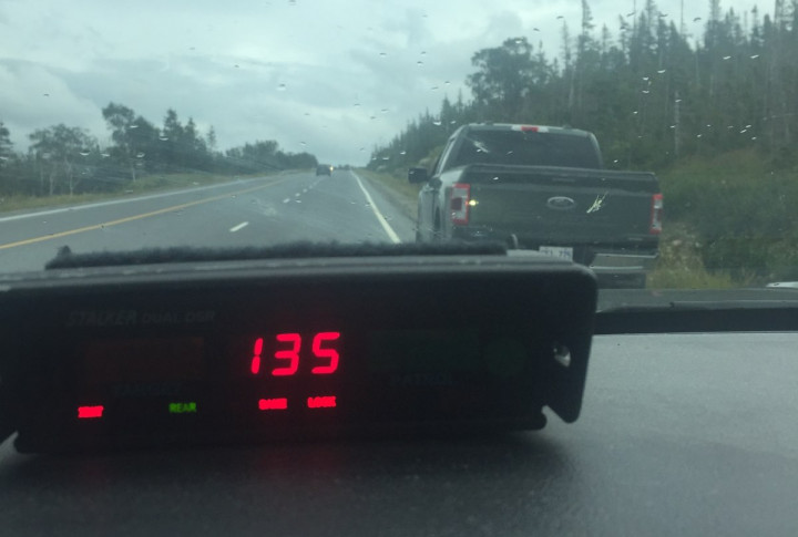 Le radar d'une voiture de patrouille indique une vitesse de 135 km/h, en rouge. Par le pare-brise, on voit la route et une camionnette verte rangée sur le côté; le ciel est couvert et une pluie légère laisse des gouttes sur le pare-brise.