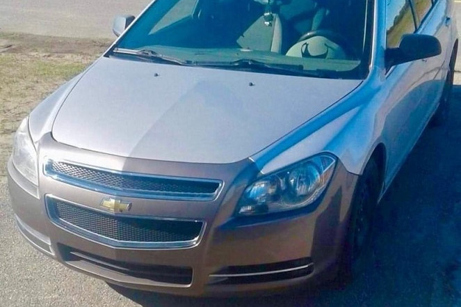 The front view of an unoccupied 4-door 2009 grey Chevrolet Mailbu is pictured parked on asphalt. The car has a brown bumper. The is an intersection in the background.