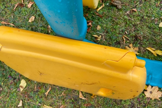 A yellow plastic door belonging to a plastic play structure is pictured with a crack in the hinge. It rests up against a blue portion of the play structure.