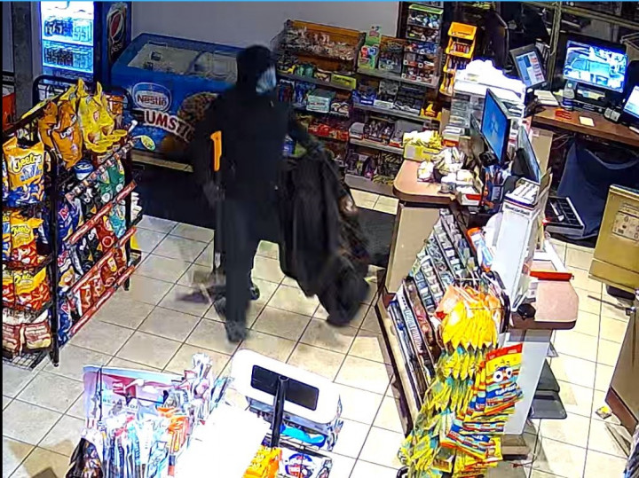 A masked individual wearing a black hoodie, a face mask, dark colored gloves and dark colored pants is captured on surveillance inside a convenience store. The individual is holding an axe in one hand and a dark colored duffle bag in the other hand.