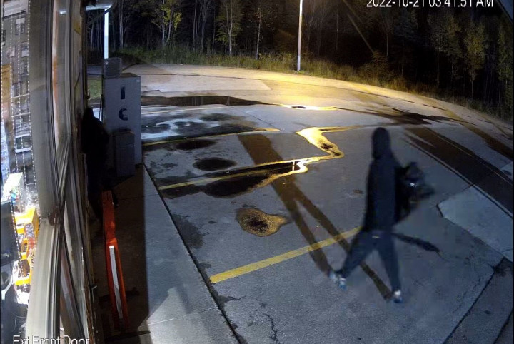 An individual wearing a black hoodie and dark colored pants is walking across the parking lot of a gas station at night during the time of a break and enter. A dark colored duffle bag is draped over the individual's right shoulder.